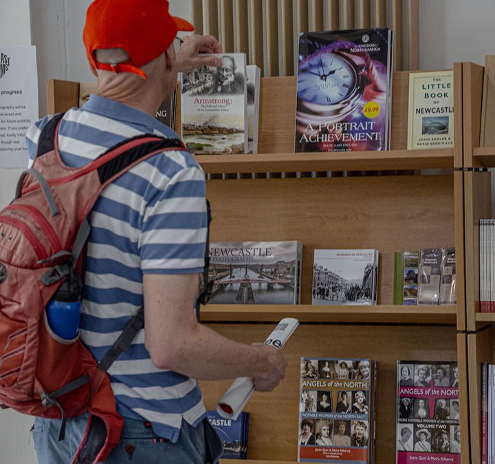 Man in red cap and blue and white striped t-shirt reaching to pick up a book from the shelves of books in Segedunum gift shop 