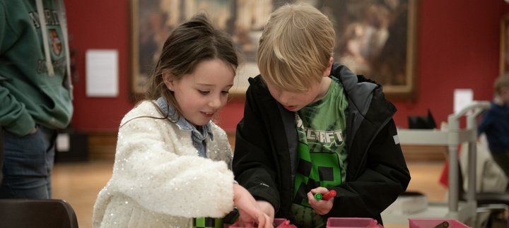 Two children looking inside a box of pens on a table, in front of a large historic painting.