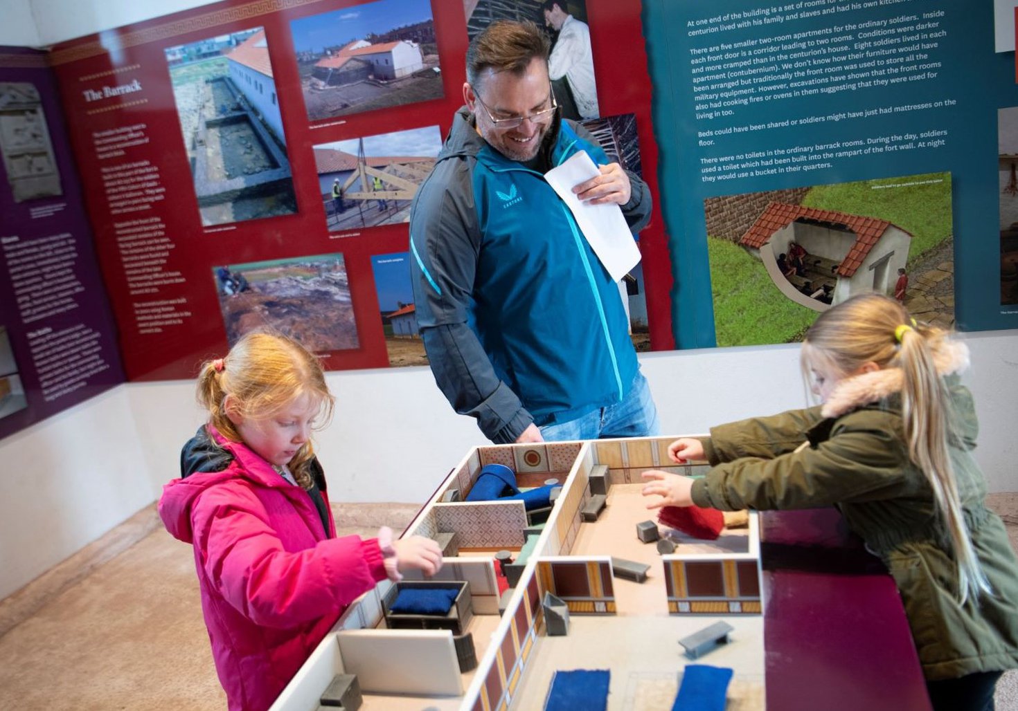 Three people, including two children and one adult, interact with a model display of the Commanding Officer's house in the reconstructed room. The background features informational panels about the fort