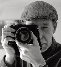 A portrait of a man wearing a flat cap, holding a camera and looking down the viewfinder. 