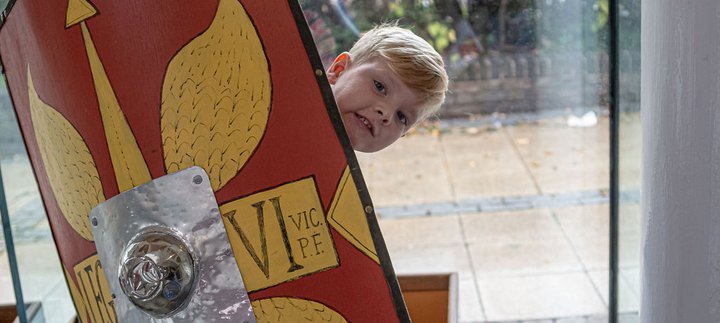 Young boy peeping out from behind a Roman style shield in the shop