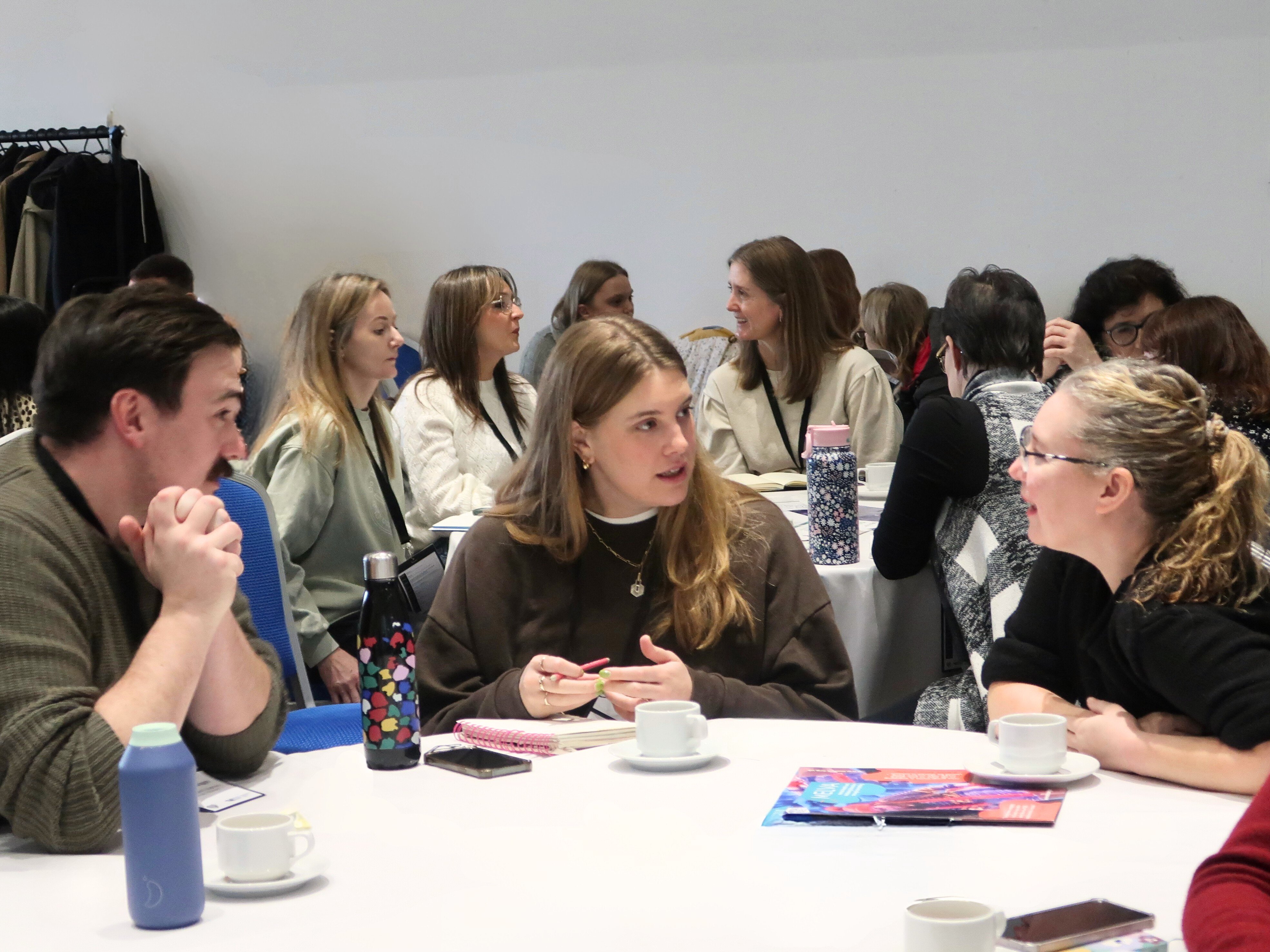 A large group of people sat in conversation at different tables at a conference. A large group of people sat in conversation at different tables at a conference.