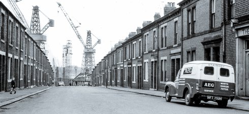 Black and white photo of street with terraced housing leading to shipbuilding yard and cranes