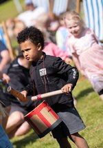 A boy with a cardboard drum around his neck, surrounded by other children playin on grass 