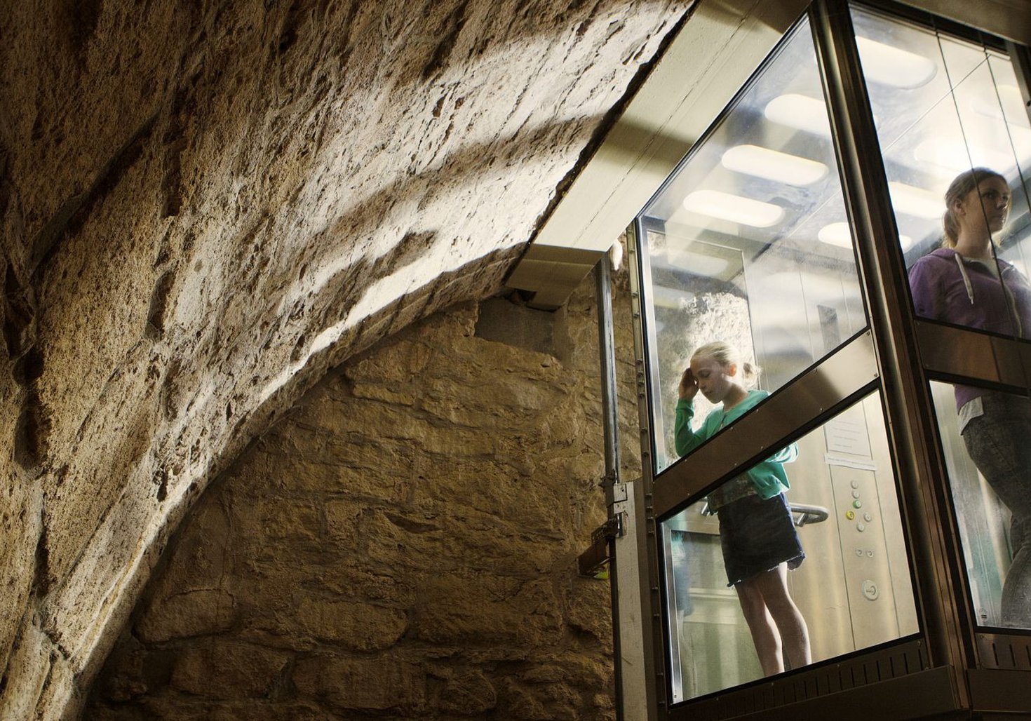Two children are in a glass lift looking out at the stone wall of Hexham Old Gaol's dungeon