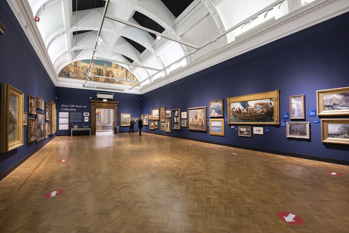A photograph showing a gallery space with dark blue walls and paintings of all different sizes and frames hung on the walls.