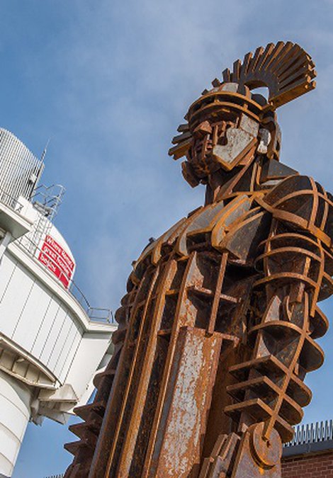 Rusted centurion sculpture with the tall glass viewing tower in the background against a blue sky