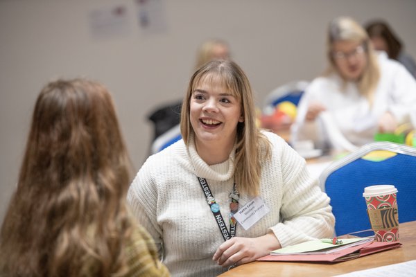 A person with blonde hair and a cream jumper, sitting at a table talking to the person next to them. A person with blonde hair and a cream jumper, sitting at a table talking to the person next to them.