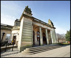 A photograph of the Shipley Art Gallery exterior. The building is made of yellow stone and the front has stone columns and two statues on the roof. 