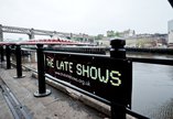 The Late Shows yellow and black banner on Gateshead quay with Swing and High Level bridges in the distance
