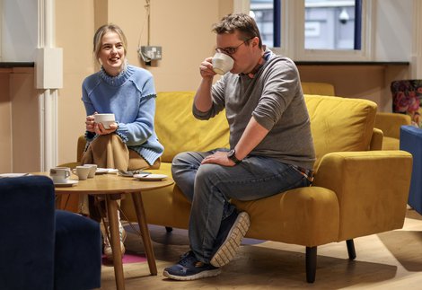 Two people sat on a yellow sofa talking and drinking