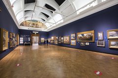 A photograph showing a gallery space with dark blue walls and paintings of all different sizes and frames hung on the walls. 