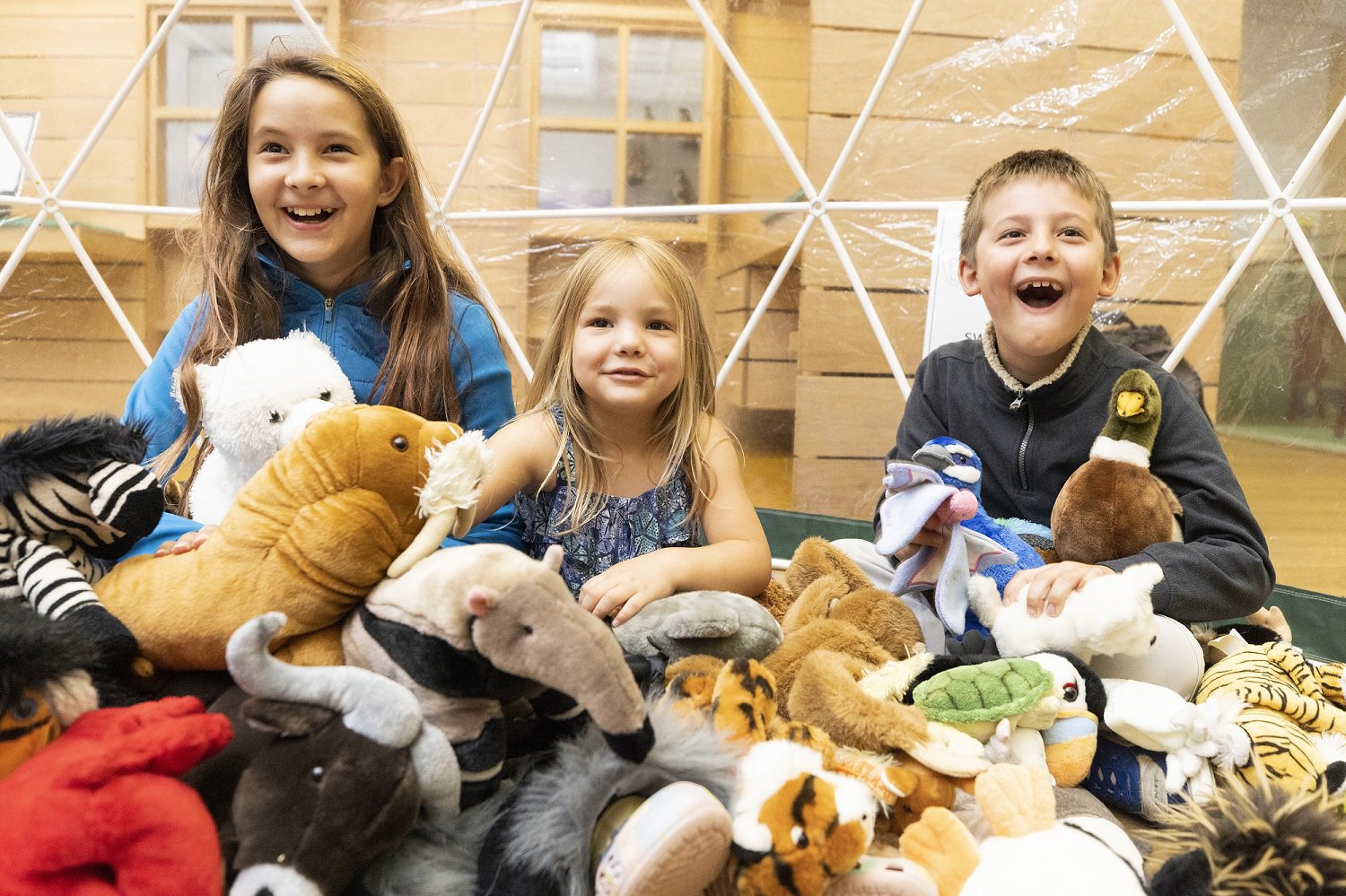 Three children at a museum surrounded by soft toys.