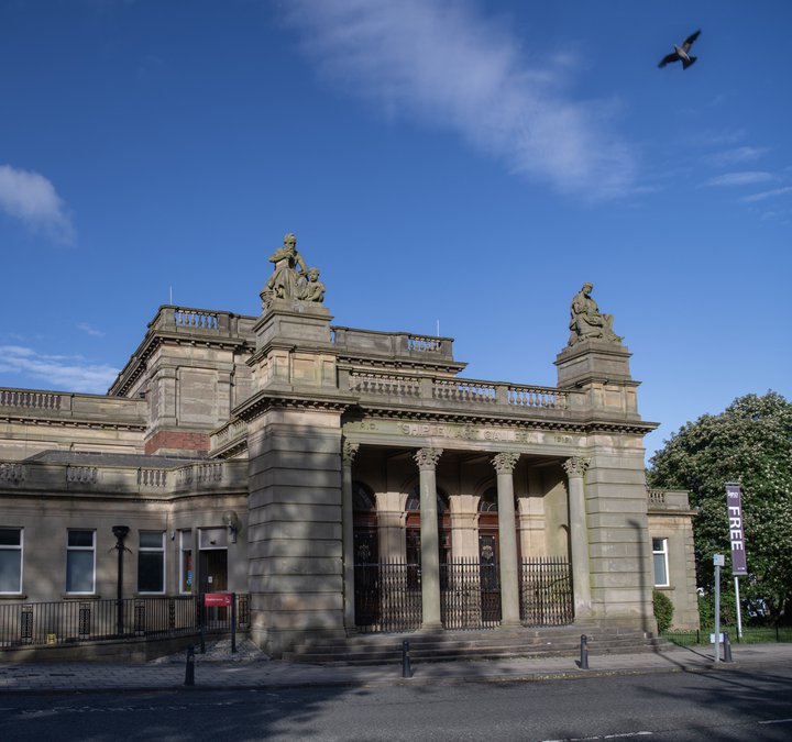 The exterior of a large stone building, with a blue sky above it. There are round columns either side of the entrance and two large pillars with statues on the top.