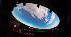 Visitors look up at a domed planetarium show. They are seeing clouds moving across a daylight sky