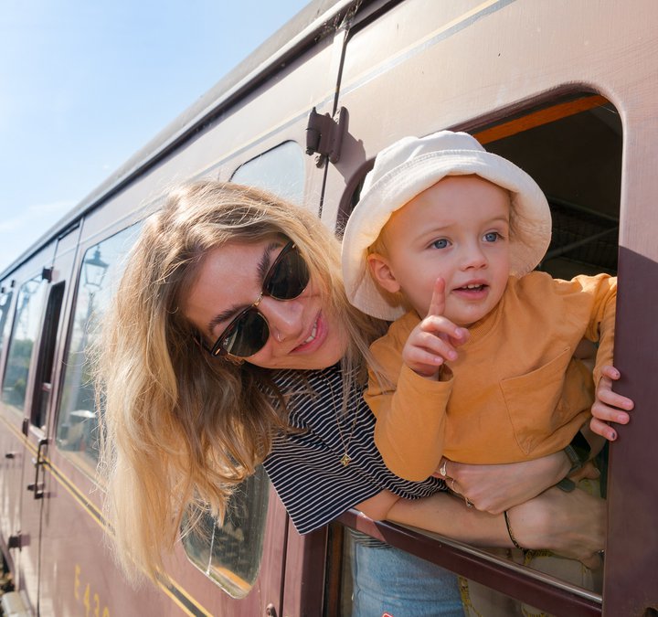 Woman with blonde hair and wearing sunglasses and her small child with mop hat leaning out of the window of a railway carriage