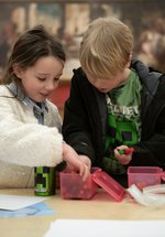 Two children looking inside a box of pens on a table, in front of a large historic painting. 
