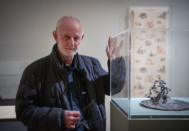 A photograph of a man posing with his sculpture, which is in a case in a gallery.