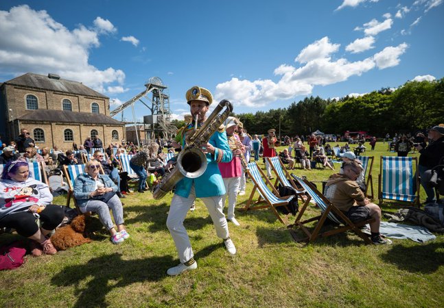 A man plays a saxophone to a group of people sitting on picnic chairs 