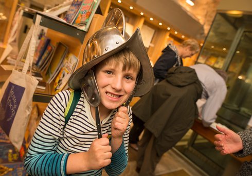 A child stands in a museum shop wearing a medieval helmet and smiling at the camera