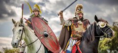 Two Roman soldiers on horseback, one with a spear and shield on a white horse, the other with a raised sword on a black horse, ready for battle under an overcast sky.