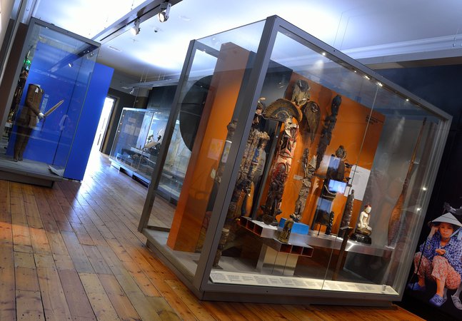 View of ethnographic items in glass museum cases. There are headdresses, statuettes and suits of armour