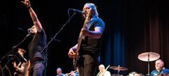 Band performing on stage with various instruments including guitar, drums, and brass.