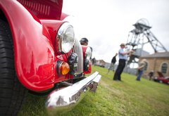 A bumper of a red vintage car parked on the grass in front of Woodhorn colliery. Men in the background are taking photographs