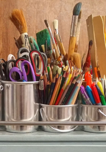 A photograph of different art materials in metal cups- pencils, paintbrushes, scissors, and more