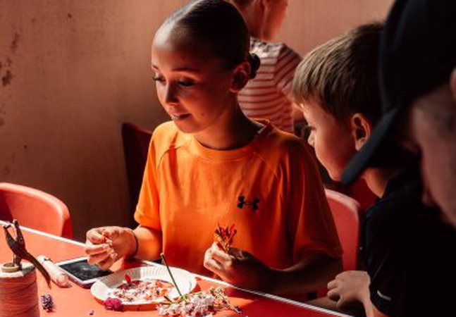 Girl in orange top sat at a craft table with others looking at the craft supplies available