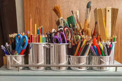 A photograph of different art materials in metal cups- pencils, paintbrushes, scissors, and more