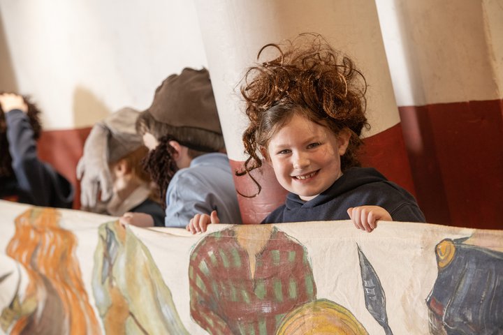 Young child wearing a wig of curly hair standing behind material painted with clothing