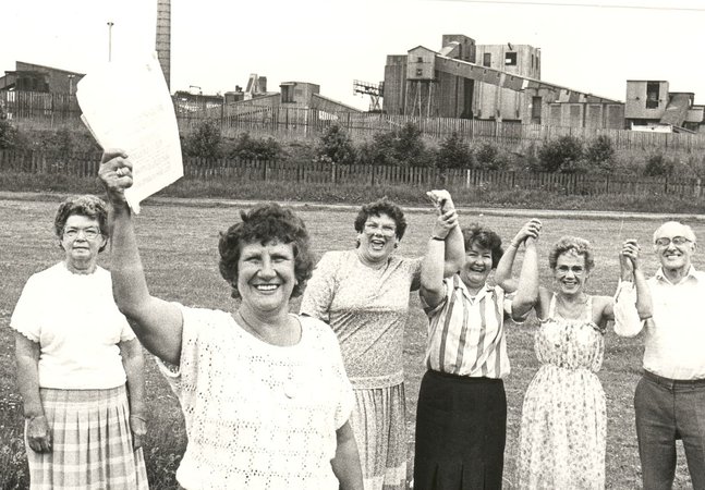 Black-and-white historical photograph of six community members standing on a grassy field in front of an industrial site. A woman in the foreground holds a document aloft, while others stand with raised joined hands. 