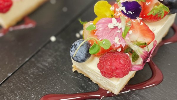 A rectangular slice of cheesecake topped with fruit and edible petals on a slate plate A rectangular slice of cheesecake topped with fruit and edible petals on a slate plate