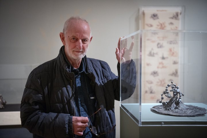 A photograph of a man posing with his sculpture, which is in a case in a gallery.