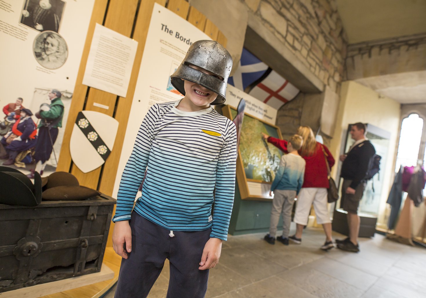 A child stands in the middle of the Milburn Room at Hexham Old Gaol, they are wearing a medieval helmet. In the background is a family looking at the display