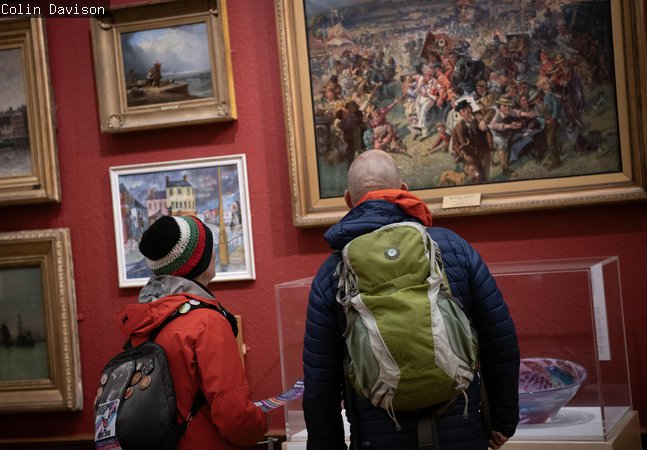 Two visitors standing and looking at a painting on a red gallery wall.