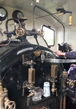A young person wearing a cap and overalls standing in front of rods and levers on the foot plate of a steam train
