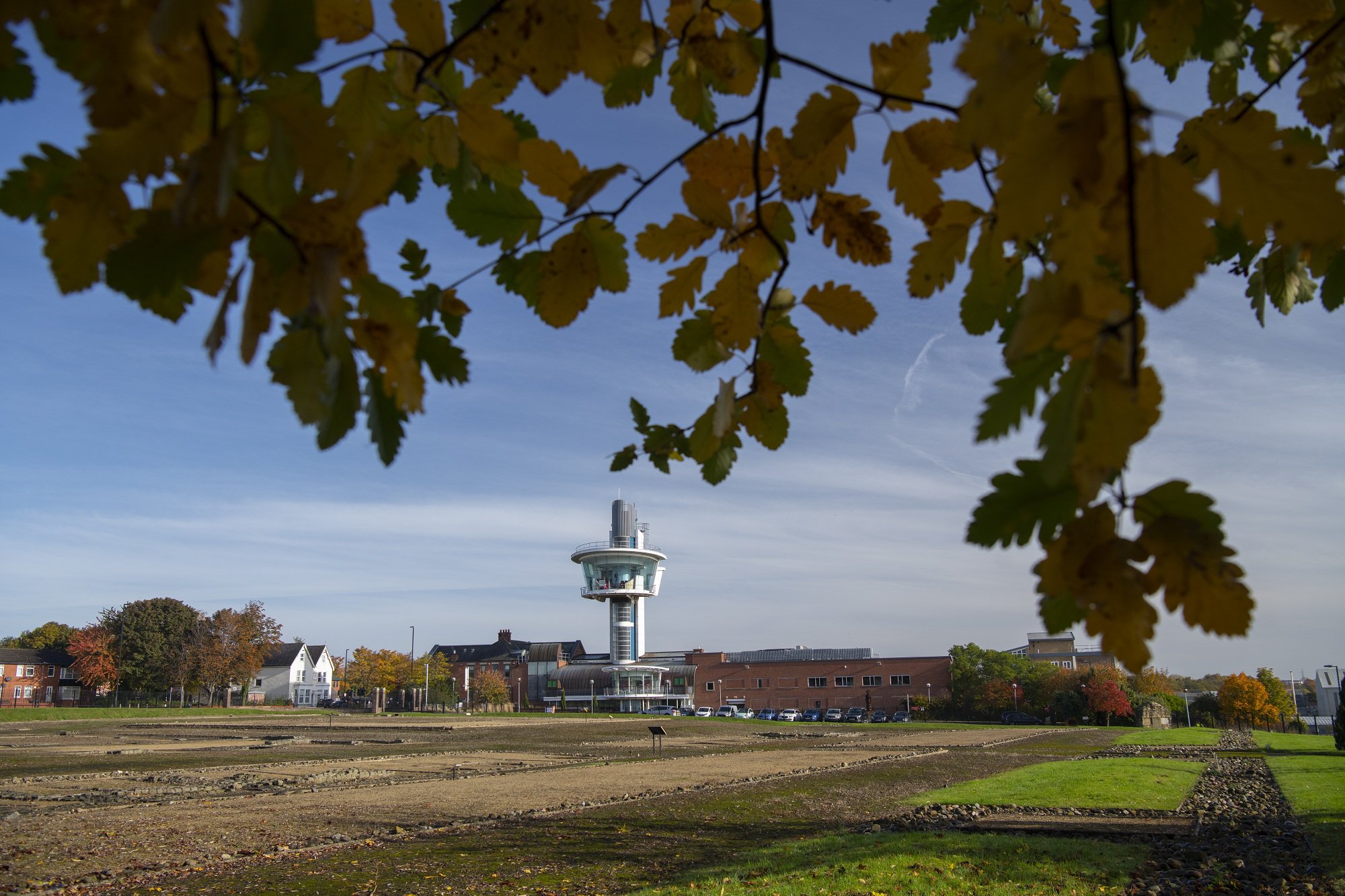 A large gravelled area leads to a modern viewing tower and brick buildings in the distance. Tree leaves hang down in the foreground. A large gravelled area leads to a modern viewing tower and brick buildings in the distance. Tree leaves hang down in the foreground.