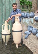 Man standing next to two amphorae beside a collection of different types of clay pots