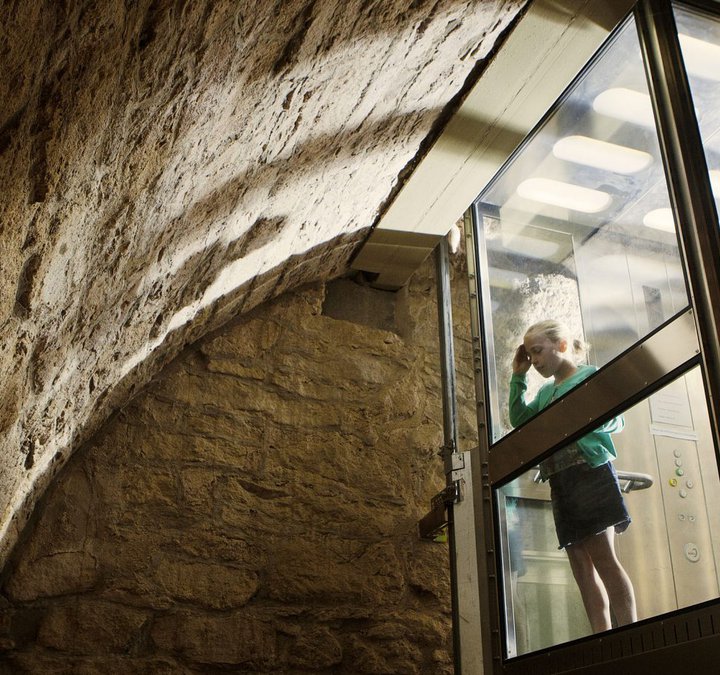 Two children are in a glass lift looking out at the stone wall of Hexham Old Gaol's dungeon