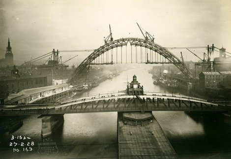 A river with a low bridge with turret in the foreground and a larged arched bridge being built using cranes in the background