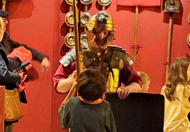 Roman solider holding a spear and shield in Shrine of the Standards talking to a young boy
