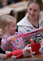 A photograph of a mum and her toddler crafting. The toddler holds a pink metallic ribbon.