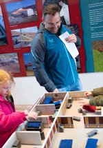 Three people, including two children and one adult, interact with a model display of the Commanding Officer's house in the reconstructed room. The background features informational panels about the fort
