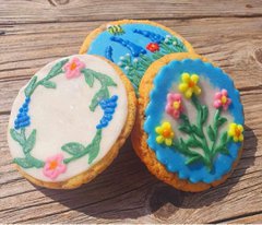 A photograph of three cookies decorated with floral designs. The flowers are pink, yellow, and blue with green stems.