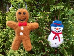 A needle felted gingerbread man next to a needle felted snowman hung on a conifer tree.