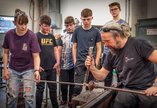 A group of students wearing eye protection gather round an artist doing glass blowing 