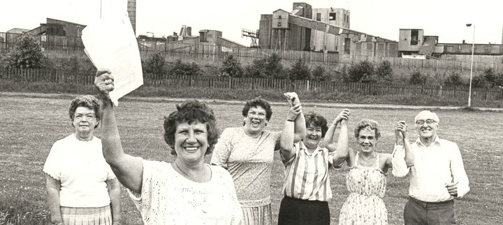 Black-and-white historical photograph of six community members standing on a grassy field in front of an industrial site. A woman in the foreground holds a document aloft, while others stand with raised joined hands. 