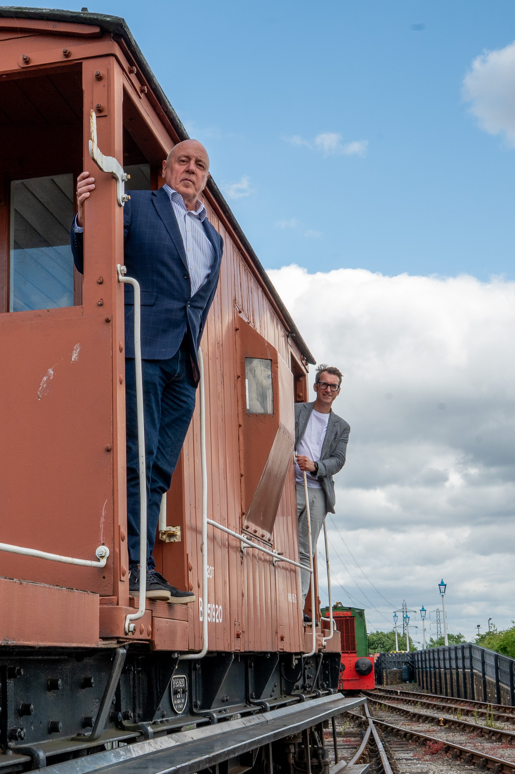 Two men are stood leaning out of a stationary railway vehicle, looking towards the camera.